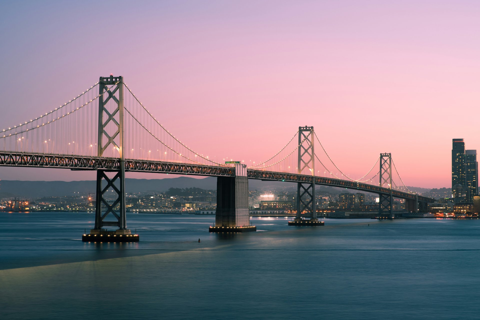 A suspension bridge spans across calm water at dusk, with city lights twinkling beneath a pink and purple sky, evoking a serene and majestic atmosphere