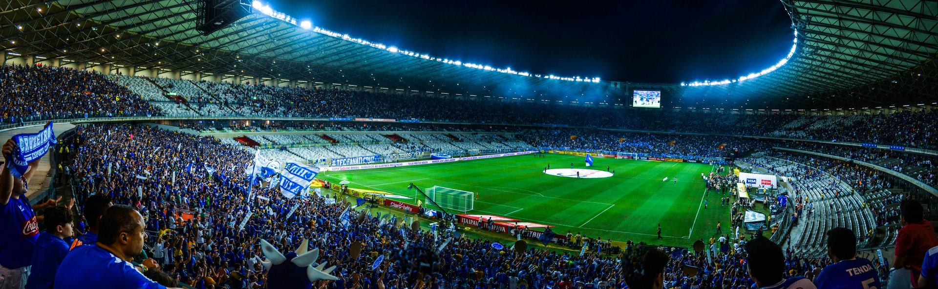 A panoramic view of a large soccer stadium filled with fans, with bright stadium lights illuminating the green field and players preparing for a match.