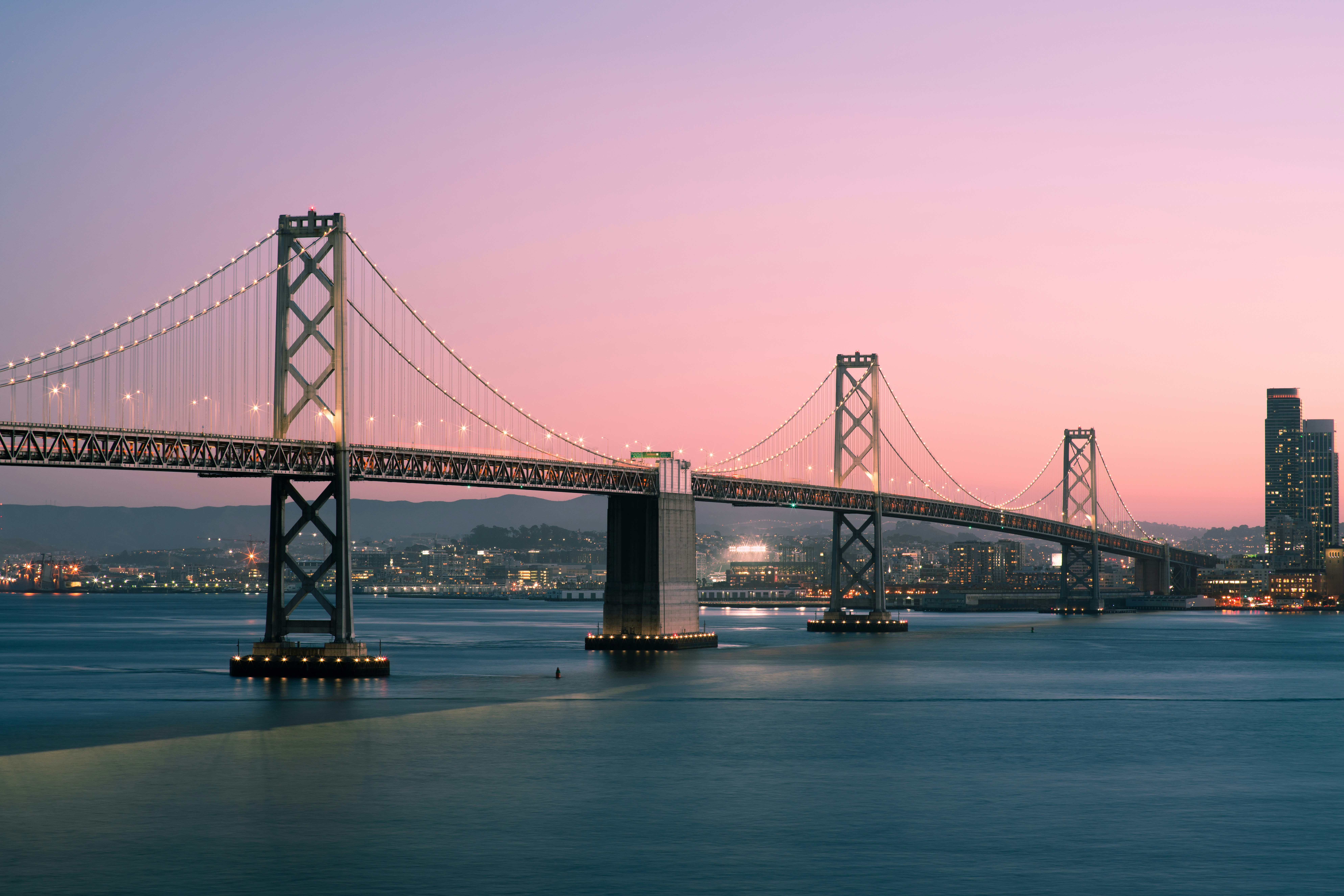 A suspension bridge spans across calm water at dusk, with city lights twinkling beneath a pink and purple sky, evoking a serene and majestic atmosphere